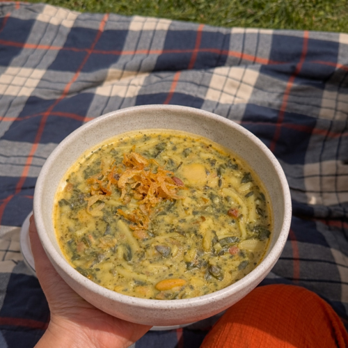 A bowl of Persian Ash Reshteh soup topped with mint oil, fried onions, and creamy kashk, filled with herbs, beans, lentils, and noodles beside a fresh melon margarita cocktail.