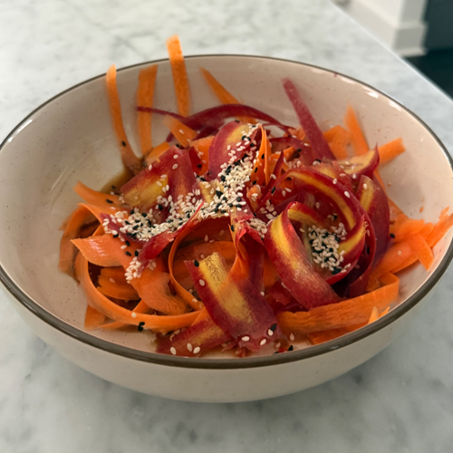 A vibrant carrot salad made with ribboned carrots, sesame seeds, cilantro, green onions, and crushed nuts in a glossy soy-ginger dressing, served alongside an orange blossom martini garnish.